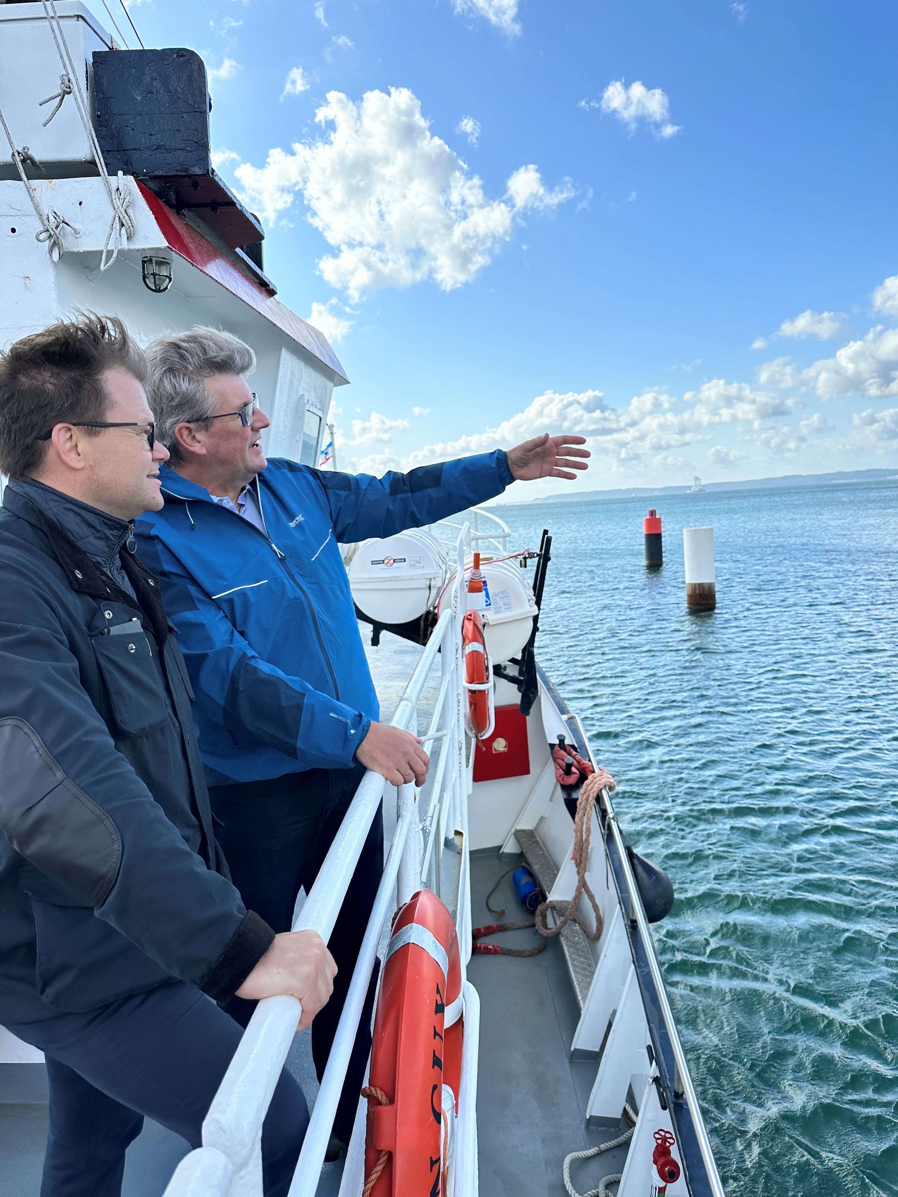 Staatsminister Carsten Schneider steht an Deck eines Schiffs und lässt sich die Begebenheiten des Hafens Mukran erklären